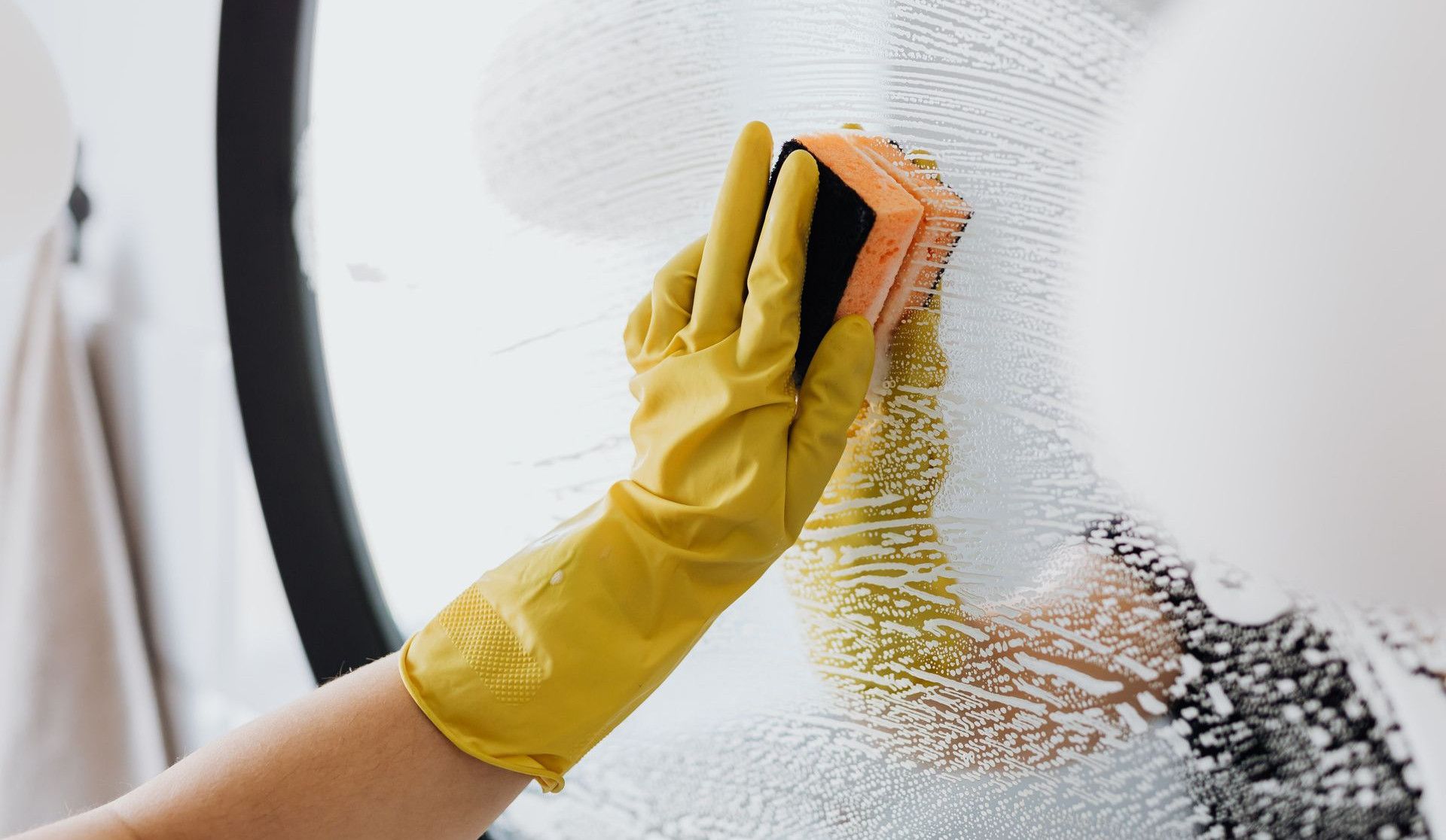 Anonymous person in yellow rubber gloves using simple sponge for cleaning mirror in bathroom